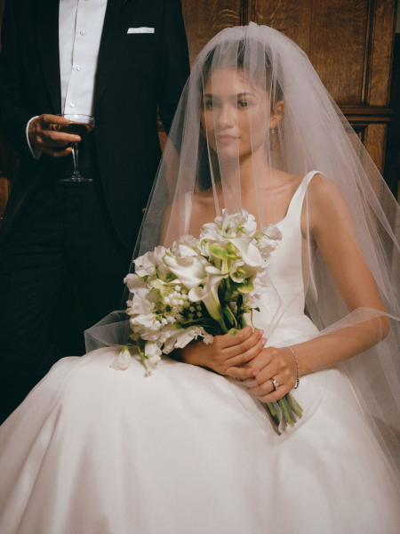 Zendaya sits dressed in a wedding gown and veil holding a bouquet of flowers.